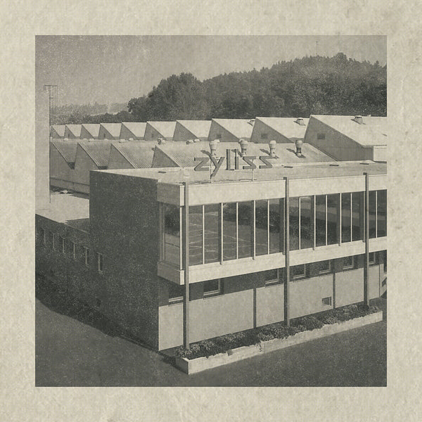 A black and white photo of a mid-century modern industrial building with large windows and a roof of repeating triangular peaks. The sign Zyliss is displayed on top, and trees are visible in the background.