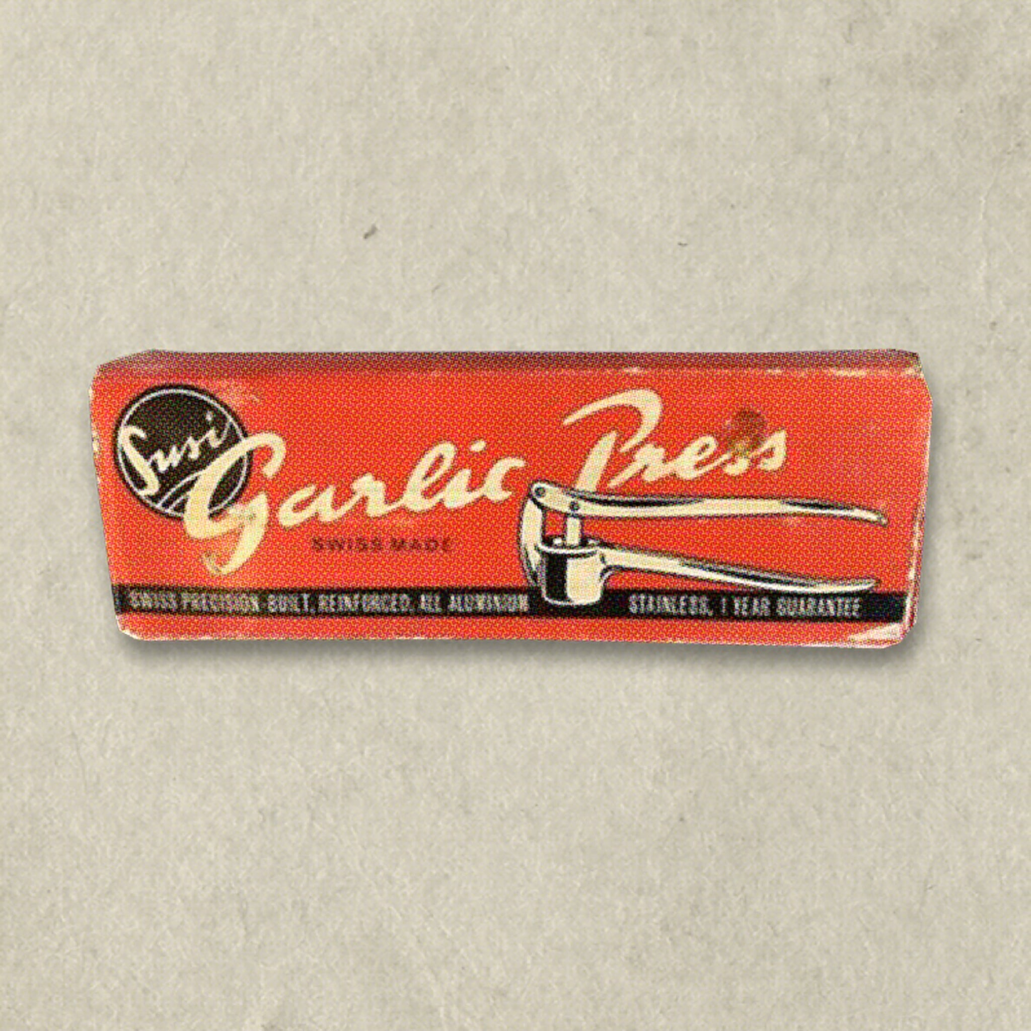 A vintage red and black box labeled Garlic Press with an illustration of a garlic press tool and the words Swiss made on a light, textured background.