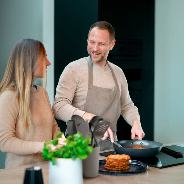 A man in an apron cooks at a stove with the Zyliss Turner XL, smiling at a woman beside him. They chat in a modern kitchen filled with herbs and utensils featuring ergonomic handles.