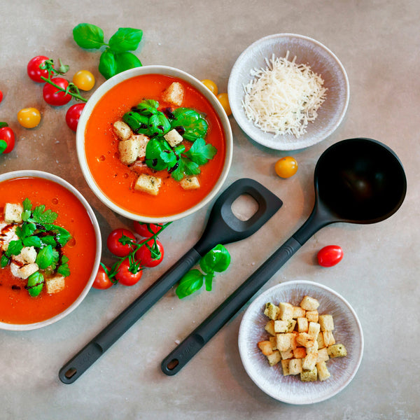Two bowls of tomato soup topped with croutons and fresh herbs, surrounded by cherry tomatoes, a small bowl of grated cheese, a bowl of croutons, and two black serving utensils on a light surface.